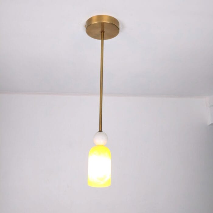 A lit Modern Alabaster Pendant Light hanging from a ceiling. The fixture features a round brass canopy, a sleek gold-finished downrod, and a glowing cylindrical shade crafted from natural white and yellow translucent alabaster stone.